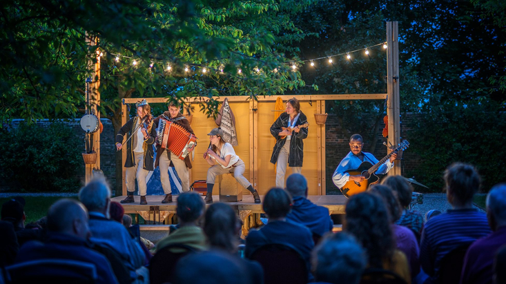 Five actors are playing music instruments up on stage as part of a evening open air performance.